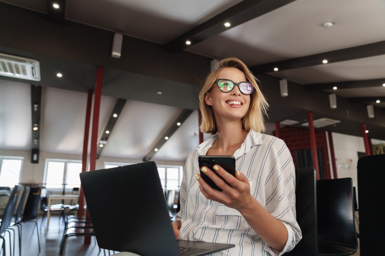 Photo of laughing young woman wearing eyeglasses working on laptop and cellphone while sitting in conference hall